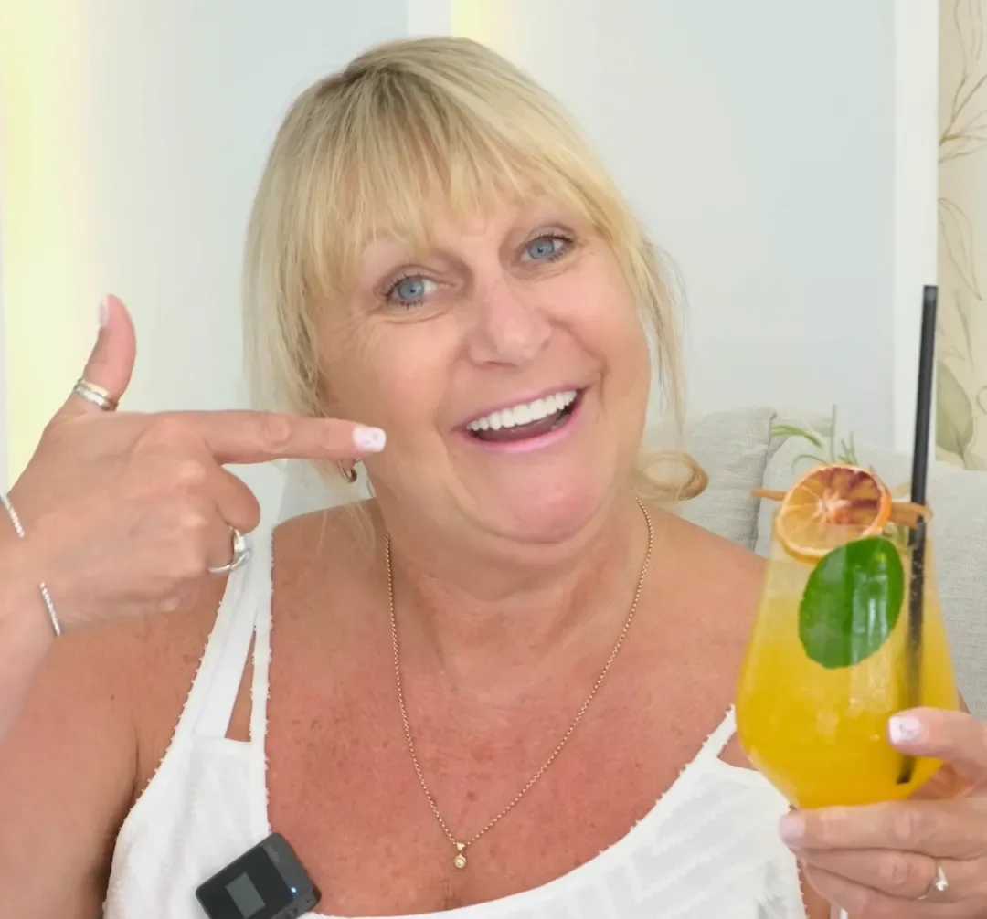 A smiling UK Smiles patient pointing to her new teeth with a drink in hand