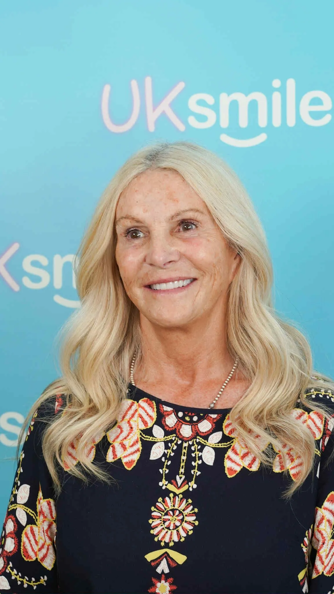 Older blonde woman smiling in front of the UK Smiles backdrop