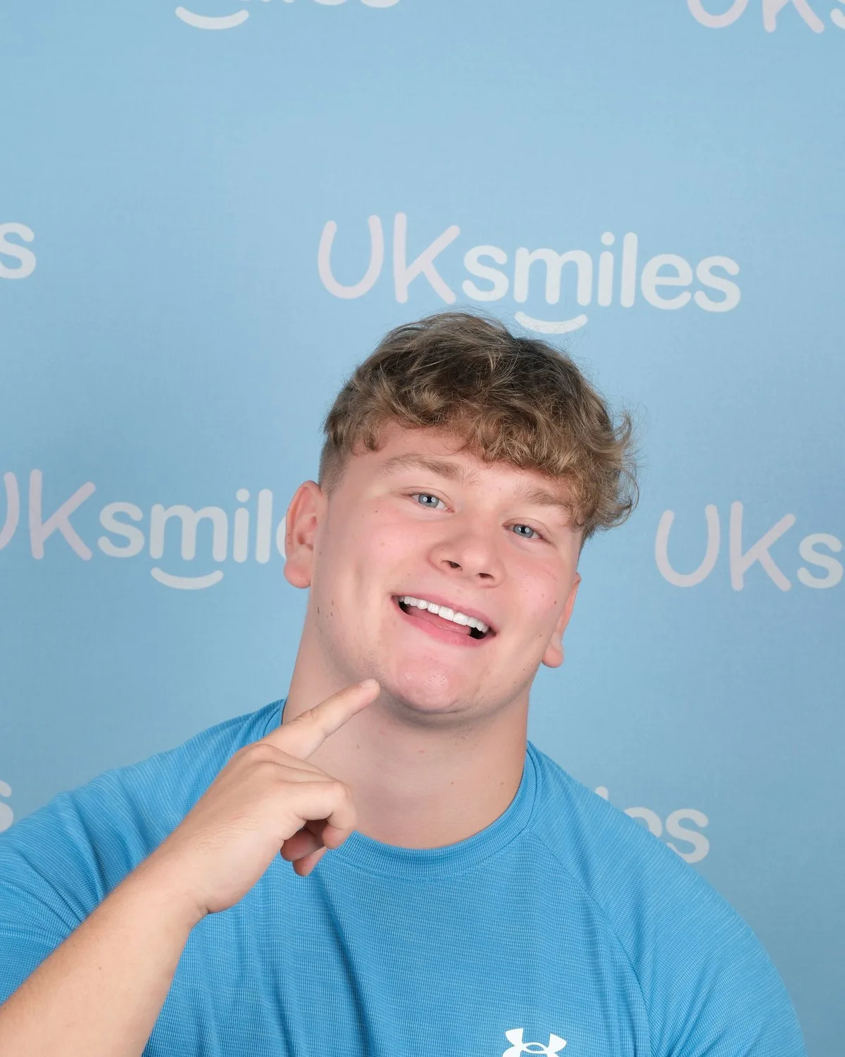 UK Smiles patient smiling after a full set of Emax veneers, photographed in front of the cyan UK Smiles backdrop in Antalya.